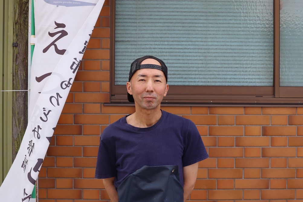 左側には「えつ」と書かれた白い旗がたなびき、レンガの壁と窓を背景に、紺色のTシャツと黒いキャップを着用した吉田宏毅さんが、微笑みながら正面を向いて立っている
