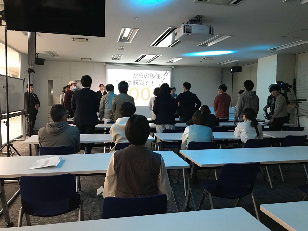 明るい照明の会議室にて、前方のスクリーンに映る「移住や転職」に関するスライドを、デスクに座る参加者や立っているスーツ姿の男女など十数名が注視しており、室内には撮影用のカメラや照明機材も配置されている写真