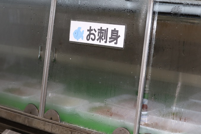 表面に水滴がつき白く曇ったガラス扉の冷蔵ショーケースに、水色の魚のイラストと共に黒い文字で「お刺身」と書かれた白いラベルが貼られ、内部に商品が並んでいる様子を斜め前から捉えた写真