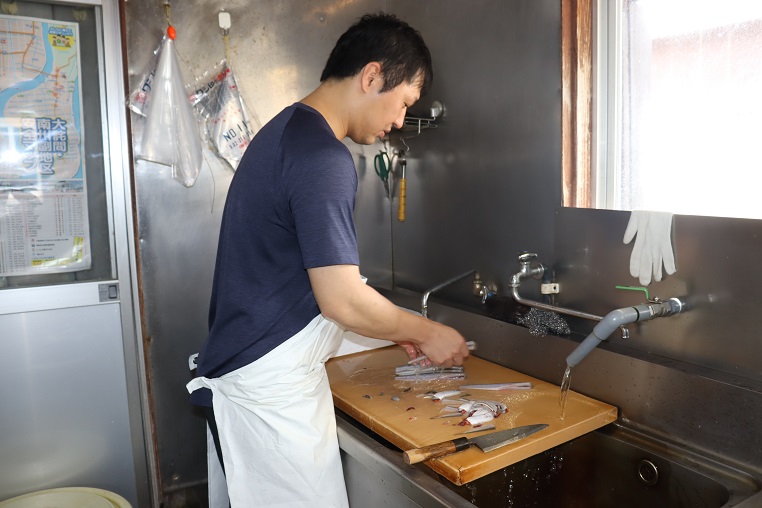 調理場で、紺色の半袖シャツに白いエプロンを着用した廣重明彦さんが、木製のまな板の上で銀色の魚を包丁で捌き、蛇口から流れる水で洗い流しながら作業に集中している横顔の写真