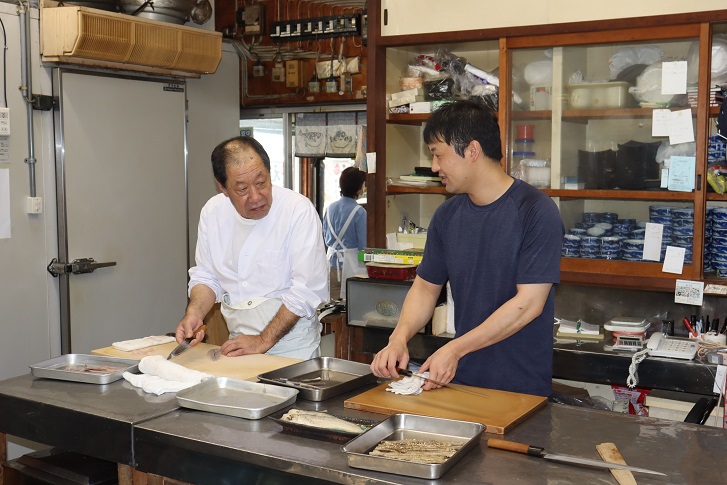 調理道具や食器が並ぶ棚を背景にした作業場で、割烹着を着た廣重寿弘さんと紺色のシャツに白いエプロンをつけた廣重明彦さんが、それぞれ包丁を手にまな板の上の魚を捌きながら、視線を交わして会話をしている調理風景の写真