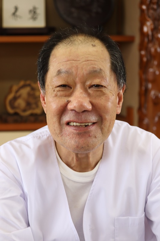 白い丸首シャツにVネックの割烹着を重ねて着用した廣重寿弘さんが、正面を向きながら穏やかに歯を見せて微笑んでいる明るい色調の上半身の写真