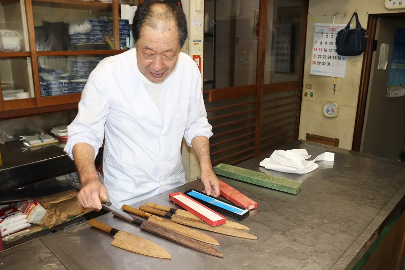 調理場で、白い割烹着を着た廣重寿弘さんが、調理台の上に並べられた長さや形状の異なる複数の和包丁や木製の鞘、砥石を指し示しながら、それらの道具について説明している様子を捉えた写真