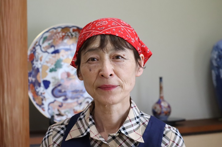 赤いバンダナを頭に巻き、チェック柄のシャツと紺色のエプロンを身に着けた店主の女性が、背後に大きな絵皿が飾られた和風の室内で穏やかな微笑みを浮かべながら正面を見つめている上半身の写真