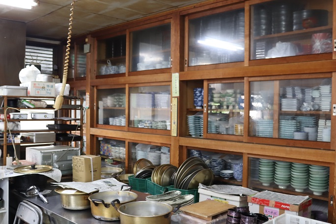 木製の大きな棚に整然と積み重ねられた大量の和食器や、調理台の上に置かれた金色の鍋や蓋、段ボール箱などが並び、奥には業務用電子レンジも見える、昭和レトロな雰囲気が漂う広々とした調理場の写真