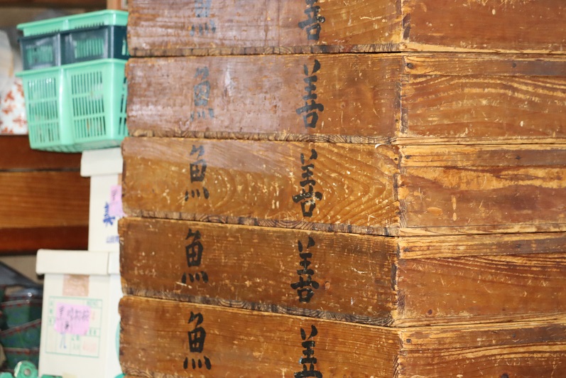 使い込まれて年季の入った木製の番重が垂直に高く積み重ねられ、その側面には黒い墨文字で「魚善」と力強く記されており、背景には白い段ボール箱や水色のプラスチックカゴが置かれた、活気ある調理場の舞台裏を感じさせる写真