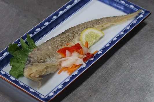 青い縁取りのある長方形の白い皿に盛り付けられた一匹丸ごとの魚の唐揚げに、彩りとしてスライスされたレモンや赤パプリカ、玉ねぎ、人参のマリネと緑の葉野菜が添えられている、シンプルで清潔感のある料理写真