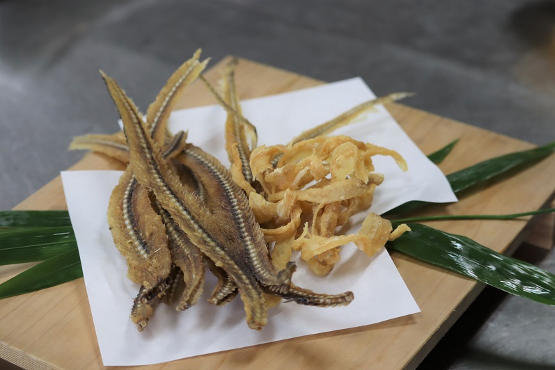 木製プレートの上に敷かれた白い和紙の上に、香ばしく揚げられた数本の魚の骨煎餅と身のフリッターが盛り付けられ、脇には鮮やかな緑色の笹の葉が添えられて素材の質感が強調されている、職人技が光る和食の揚げ物写真