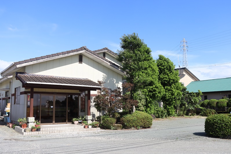 抜けるような青空の下に佇む平屋建ての和風建築で、瓦屋根の玄関アプローチの横には手入れされた庭木や植栽が並び、広々とした砂利の敷地や遠くに見える鉄塔がのどかな雰囲気を感じさせる、落ち着いた建物外観の写真