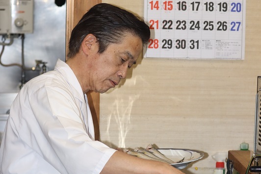 白い調理服を着た御厨和秀さんが、カレンダーが貼られた厨房の中で真剣な表情を浮かべ、平皿に盛り付けられた新鮮なエツを丁寧に調理しようとしている様子を横から捉えた写真