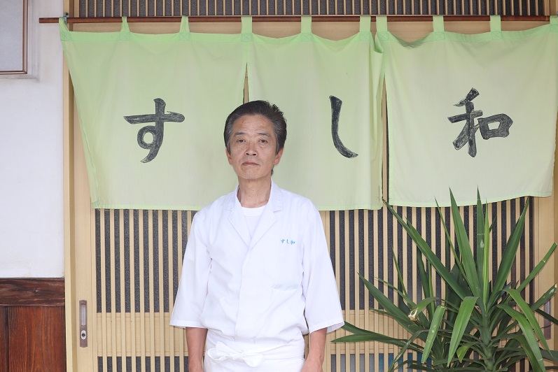 背景には格子状の引き戸と観葉植物が配置された、「すし和」という店名が書かれた薄緑色の暖簾の前の寿司屋の店先で、白い調理服を着た御厨 和秀さんが真剣な表情で立っている写真