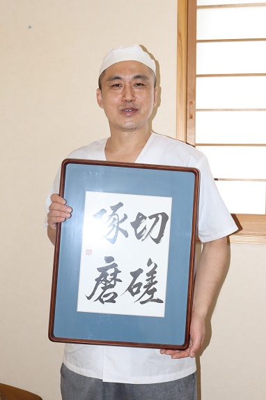 和室のような背景の前で、白い調理服と帽子を着用した店主の男性が、四字熟語の「切磋琢磨」と書かれた書道の額縁を両手で正面に掲げて持ち、柔らかな表情でカメラを見つめている写真
