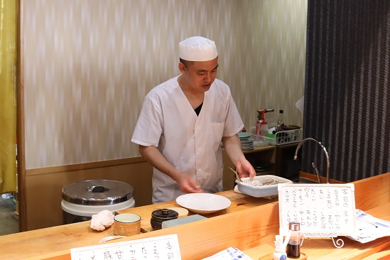 白の調理服と帽子を身に着けた料理人が、木製のカウンター越しに魚の入った白いトレイを手に持ち、手元の白い皿に向かって調理の準備を進めている様子を捉えた、店内の品書きや調理器具が並ぶ厨房の風景写真