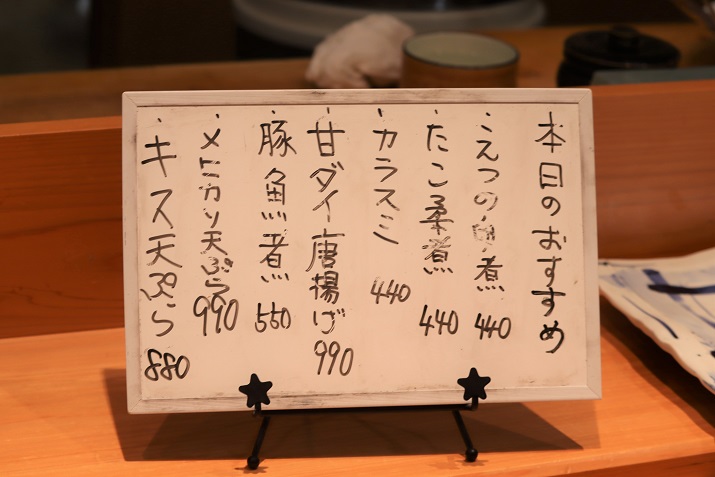 木のカウンターに置かれた白いボードに、手書きで「本日のおすすめ」と題して「えつの白煮」や「甘ダイ唐揚げ」といった品名と価格が並び、星形の留め具が付いた小さなスタンドで支えられている、店内の活気を伝えるお品書きの接写写真