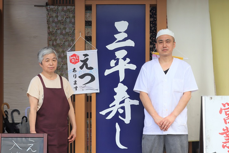 「三平寿し」と書かれた紺色の大きな看板を挟んで、白い調理服姿の男性店主と茶色のエプロンをつけた女性が店先に並んで立ち、穏やかな表情で正面を見据えている、情緒ある和食店の入り口付近を捉えた写真