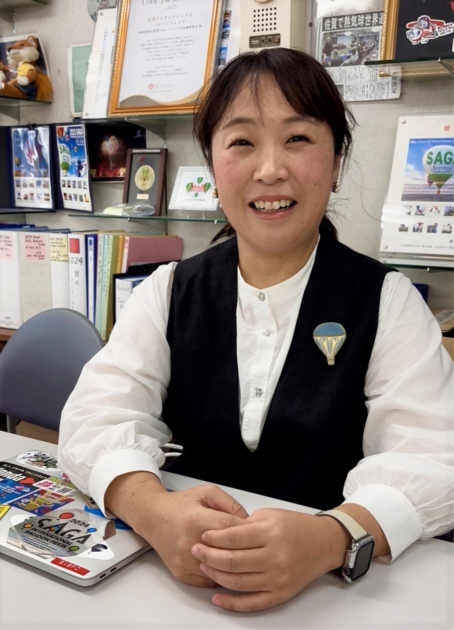 背後の棚に多くの表彰状やバルーンフェスタ関連の資料が並ぶ室内で、バルーンのブローチを胸に付けた笑顔のにしくぼまさよさんを写した写真