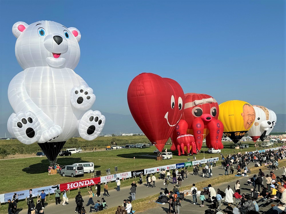 澄み渡る青空の下、広大な河川敷にシロクマやハート、タコなどの形をしたユニークな変形気球や、企業のロゴが描かれた色鮮やかな熱気球が並び、手前の通路を歩く大勢の観客と共にイベントの活気ある風景を捉えた佐賀インターナショナルバルーンフェスタの写真