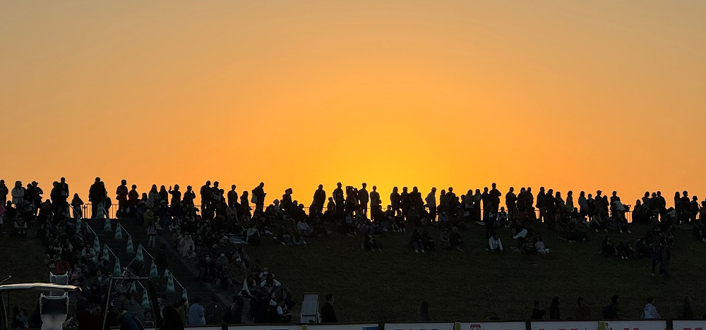 燃えるようなオレンジ色の夕焼け空を背景に、河川敷の土手で肩を並べて佇む大勢の観客たちが黒いシルエットとなって浮かび上がり、静寂の中にイベントの余韻と哀愁が漂う佐賀インターナショナルバルーンフェスタの写真