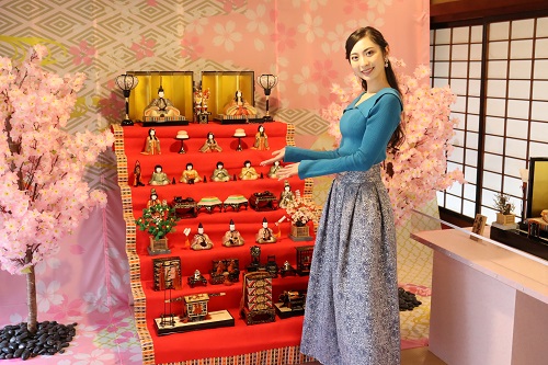 桜の花が描かれた背景の前で、青いトップスと柄物のロングスカートを着た女性が、赤毛氈が敷かれた豪華な七段飾りの雛人形を笑顔で紹介しており、周囲には満開の桜の木も配置された春らしく華やかな展示会場の写真