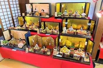 赤毛氈が敷かれたひな壇の上に、金屏風を背にしたお内裏様とお雛様の親王飾りが八組、段違いに美しく並べられており、それぞれが異なる色柄の装束を纏って和の情緒あふれる空間を華やかに彩っている雛人形の写真