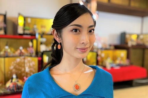 明るい青色のトップスを着た女性が、細やかな幾何学模様の佐賀錦があしらわれたオレンジ色のイヤリングとネックレスを身に着け、背景に並ぶ華やかな雛人形を背に優しく微笑んでいる、伝統工芸品とアクセサリーを組み合わせた写真