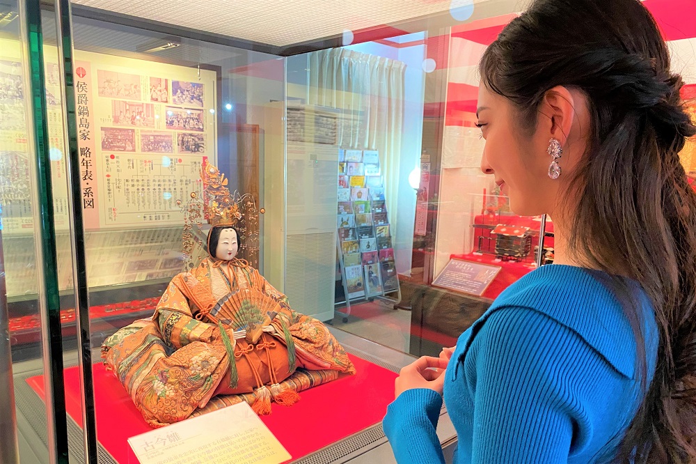 「侯爵鍋島家 略年表・系図」が展示された室内で、豪華な衣装を纏い扇を手にした古今雛を、青いトップスを着て耳飾りの光る女性が穏やかな表情で見つめている、歴史と文化の重みを感じさせる展示風景を捉えた写真