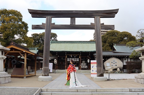 巨大な木造の鳥居の奥に緑色の屋根の社殿が建つ神社にて、鮮やかな赤や白の装束を身にまとった長い髪の女性が参道の中心で鳥居に向かって後ろ向きに佇んでいる、静謐で厳かな雰囲気が漂う神域の風景を写した写真