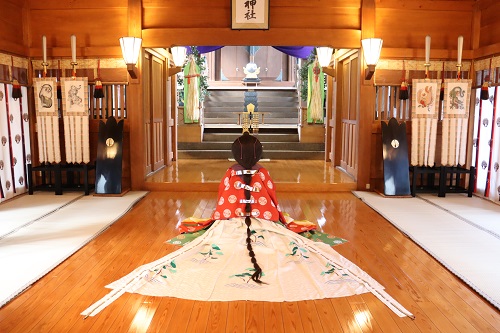木の温もりを感じる厳かな神社の拝殿内にて、赤や白の鮮やかな装束を纏い長い髪を後ろで結わえた女性が、奥にある祭壇に向かって板張りの床に静かに座り、その装束の裾が扇状に美しく広がっている様子を捉えた写真