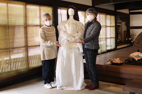 伝統的な和風建築の室内で、ウィーン万博で皇后エリザベートが着用した白いドレスを再現した衣装を中央に、制作者の南里早智子さん（左）と末岡暁美さん（右）が並び、衣装を紹介している記念写真