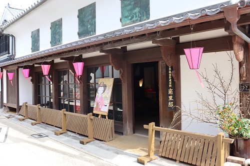 白壁に重厚な木造の梁が映える伝統的な和風建築で、軒下にはピンク色の提灯が並び、入り口には「佐賀城下ひなまつり」のポスターが掲げられた、幕末の志士ゆかりの建物である南里邸の外観写真