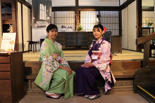 歴史ある和室の畳の間を背景に、華やかな桜柄が施された緑色と紫色の振袖をそれぞれ身に纏った二人の女性が、落ち着いた雰囲気の中で並んで座りながら穏やかな微笑みを浮かべポーズをとっている、和の情緒溢れる写真