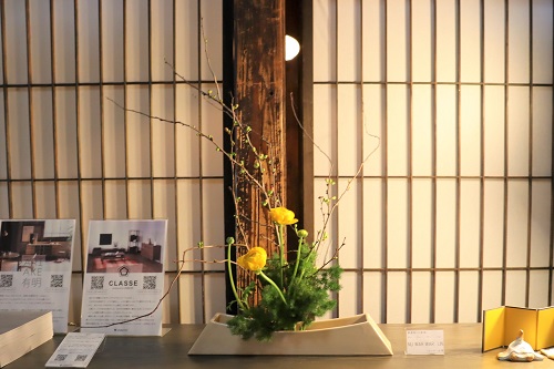 障子戸を背景に白い花器に生けられた黄色の花と繊細な枝ものの生け花が展示されている風景の写真