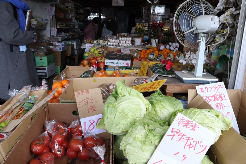 中島商店の店先で、段ボールに入ったキャベツやトマトなどの新鮮な地場野菜が並び、手書きの価格札が添えられ、奥には扇風機や果物の陳列棚、買い物をしたり接客を受けたりする人々の姿が見える活気ある商店の風景写真