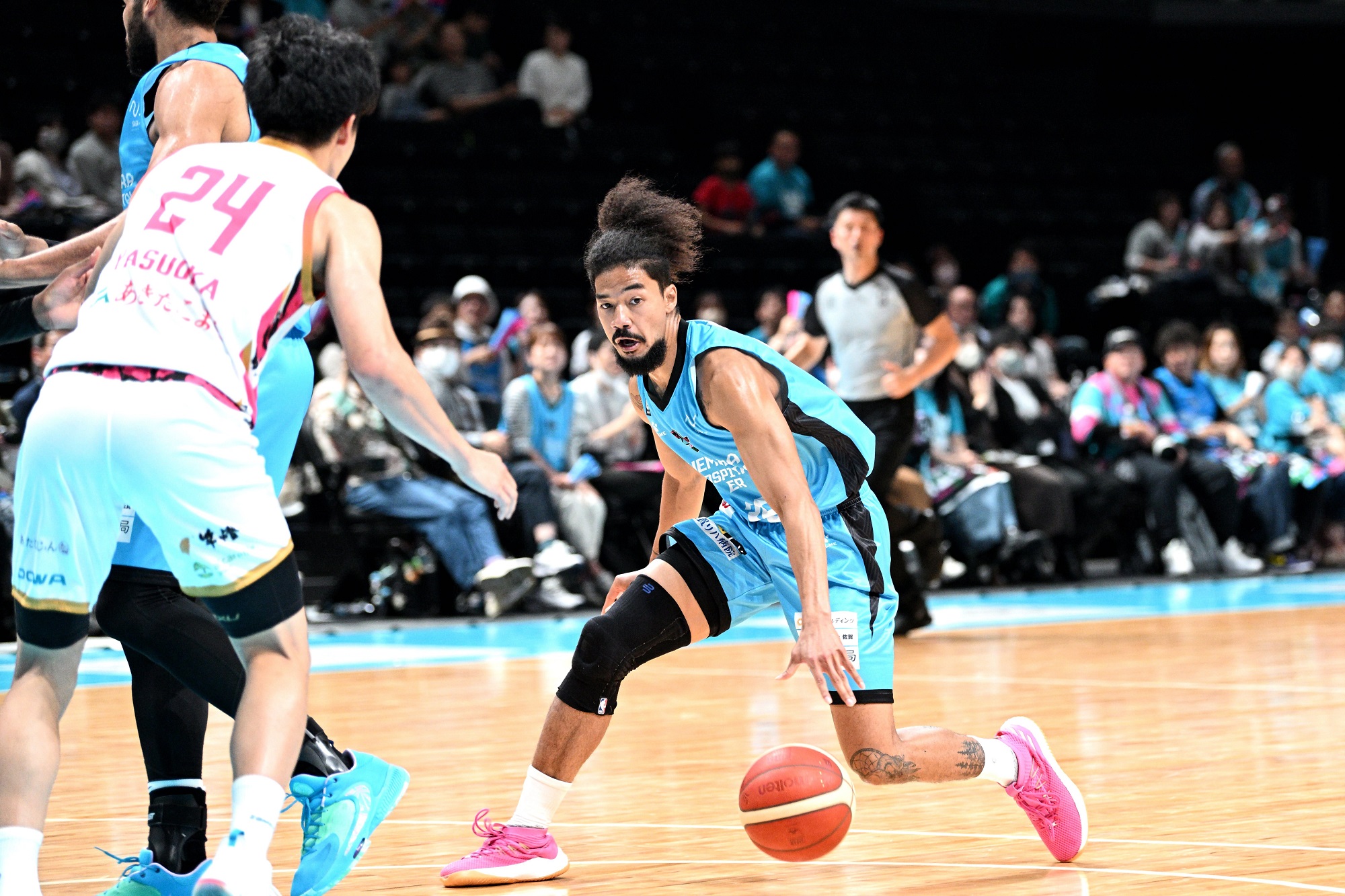 水色のユニフォームを着たアフロヘアーのバスケットボール選手が、大勢の観客が見守るコート上でドリブルを仕掛けている試合中の写真