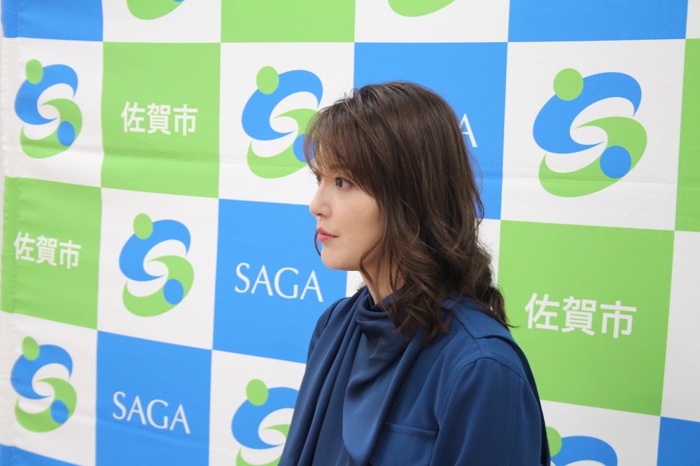「佐賀市」や「SAGA」の文字、緑と青のロゴマークが交互に配置されたバックパネルを背景に、ウェーブがかった茶髪で紺色のトップスを着た朝夏まなとさんが、物静かな表情で真横を向いている様子を捉えた写真