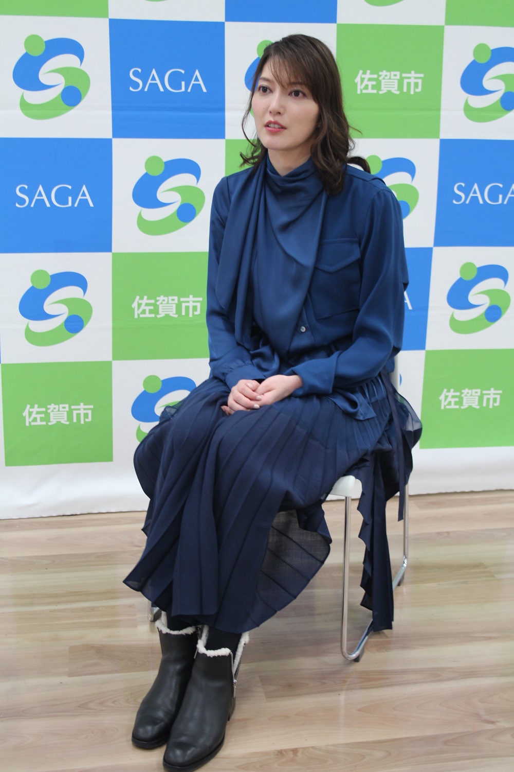 「佐賀市」や「SAGA」の文字、緑と青のロゴマークが交互に並ぶバックパネルの前に、光沢のある紺色のブラウスとプリーツスカートを纏った朝夏まなとさんが椅子に座り、斜め前を向いて静かに控えている全身の写真