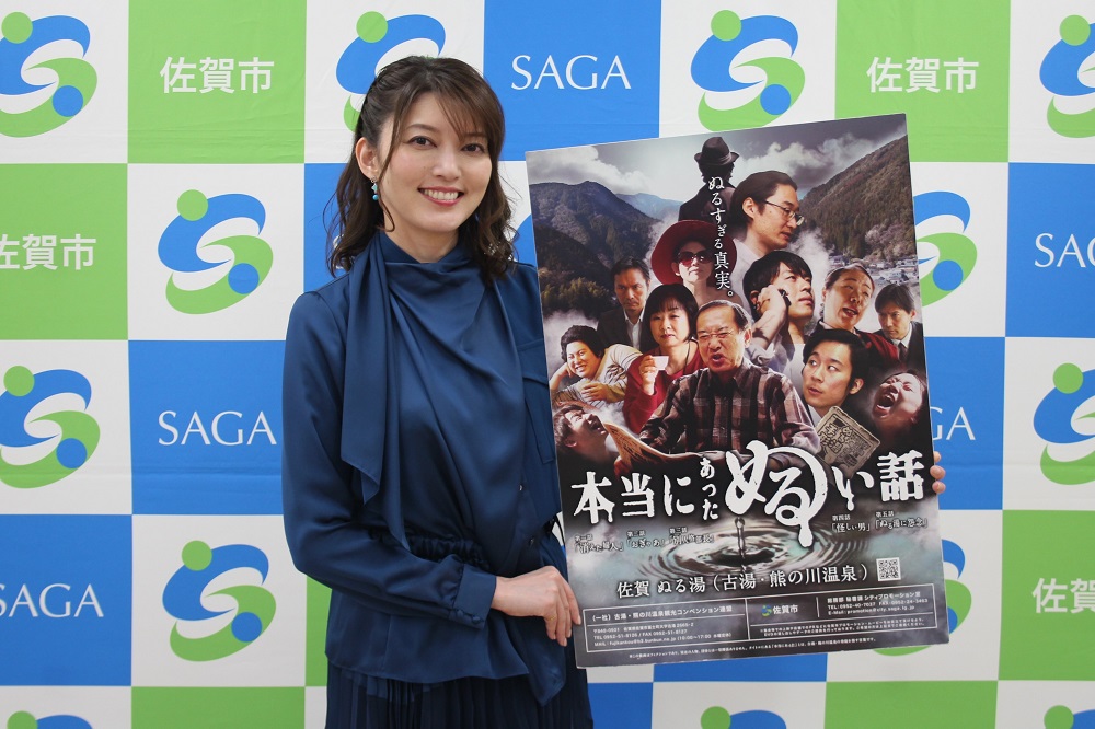 「佐賀市」や「SAGA」の文字が並ぶ背景の前で、朝夏まなとさんが「本当にあったぬるい話」と書かれた佐賀の温泉（古湯・熊の川温泉）をPRする多数の人物が写ったポスターを持ち、微笑んでいる様子を捉えた写真