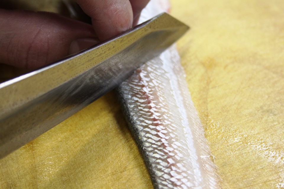木製のまな板の上で、銀色に輝く細かな鱗を持つ細長い魚の身に対し、鋭い刃物を持つ手が慎重に包丁を入れて調理を進めている様子を、質感や鱗の光沢が伝わるほどの至近距離から克明に捉えた骨切りの様子の写真