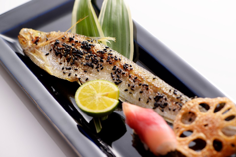 光沢のある紺色の長皿に、黒胡麻をまぶしてこんがりと焼き上げられた一匹の魚が盛り付けられ、手前には半分に切ったスダチや赤い芽生姜、レンコンの素揚げが添えられた、香ばしさが漂うような、えつの利休焼きの写真