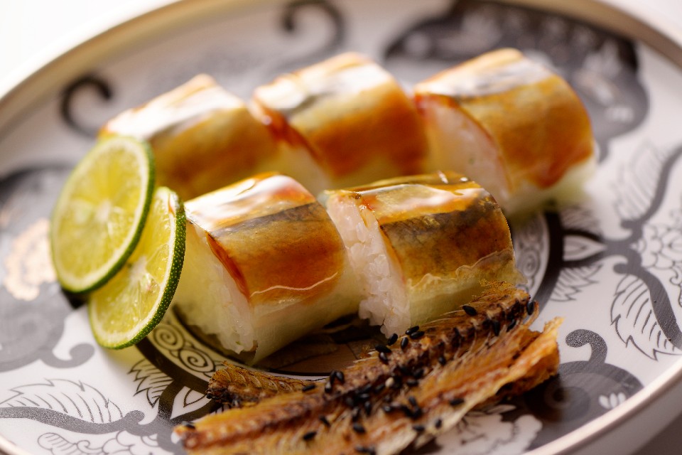 装飾的な模様が施された白い皿の上に、照り焼きにされた魚の身を乗せて美しく巻いた押し寿司が並べられ、手前には香ばしく焼かれた骨せんべいと、口直しに添えられた二枚のスダチの薄切りが彩りを添えている棒寿司の写真