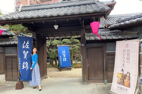 歴史ある和風建築の門の前で、青いトップスと長いスカートを身にまとった女性が佐賀錦と書かれた幟の隣に立ち、傍らには佐賀錦のおひなさまを紹介する看板が置かれた、イベントの華やかな雰囲気を伝える写真