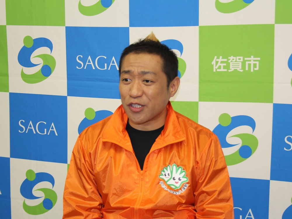 青と緑の市松模様に佐賀市のロゴが並ぶ背景の前で、胸元にキャラクターのロゴが入った鮮やかなオレンジ色のジャンパーを着たはなわさんが、口をわずかに開けて正面を見つめている上半身の写真