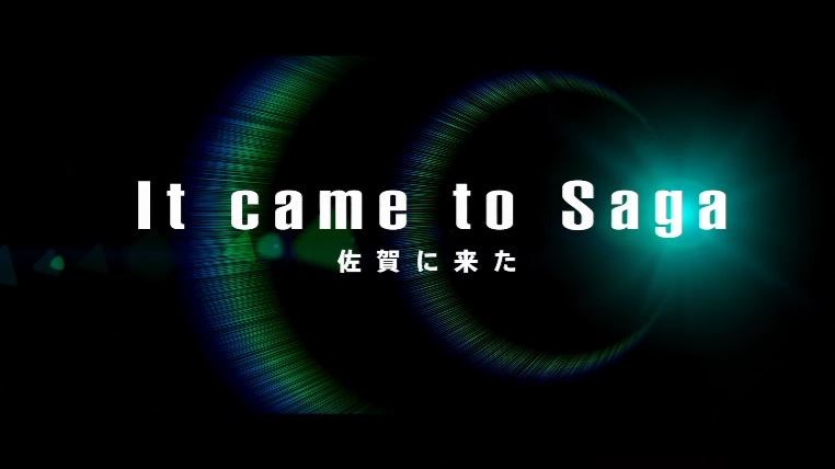 漆黒の背景に、右端から放たれる青白い鋭い光の輝きと、それを取り巻くように広がる虹色の円形レンズフレアが描かれ、中央には白抜き文字で「It came to Saga」および「佐賀に来た」と記された画像