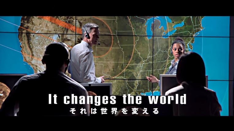 巨大なモニターに映し出された北米大陸の地図を背景に、ヘッドセットを装着した男性と女性が深刻な表情で話し合っており、手前には彼らと向き合う人物の背中と「It changes the world それは世界を変える」という字幕が添えられた写真