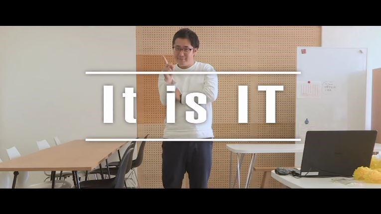 シンプルなオフィス空間で、眼鏡をかけた男性が人差し指を立てるポーズをしながら正面を向いて立っており、画面中央には上下のラインに挟まれた白抜きの太いフォントで「It is IT」という文字が大きく配置された写真