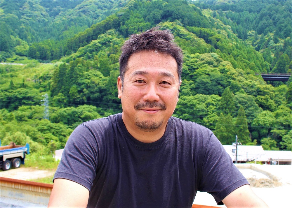緑豊かな山々が連なる背景の前で、短髪に髭を蓄えた男性が黒い半袖シャツを着用し、穏やかな微笑みを浮かべながら正面のカメラを見つめてリラックスした様子で座っている写真