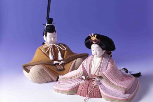 紫色の背景の前に、落ち着いた黄土色の装束を纏った男雛と、幾重にも重なる淡いピンク色の十二単が美しい女雛を並べ、優しく穏やかな表情で座る一対の雛人形の姿を捉えた写真