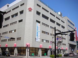 グレーのタイル貼りの外壁に赤いロゴマークが輝く佐賀玉屋の大きな建物が角地に立ち、歩道沿いにはピンク色の丸い街灯が並び、懸垂幕や道路標識が都市の活気を感じさせる百貨店の外観の写真（佐賀玉屋佐賀玉屋のサイトへリンク）