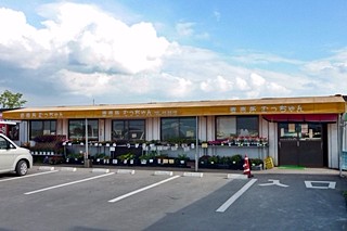 大きな入道雲が広がる空の下、黄色い長細い看板に「直売所 むっちゃん」と記された平屋の建物があり、店先の棚には多くの野菜や苗が並べられ、手前にはアスファルトの駐車場が広がる建物外観の写真