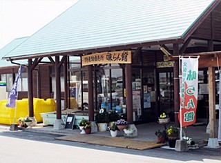 薄い緑色の大きな三角屋根と茶色の木造柱が特徴的な「久保田特産直売所 味らん館」の入り口付近を捉えており、軒下には鉢植えの花々や幟が並び、ガラス扉の向こうに店内の様子がうっすらと見える建物の写真